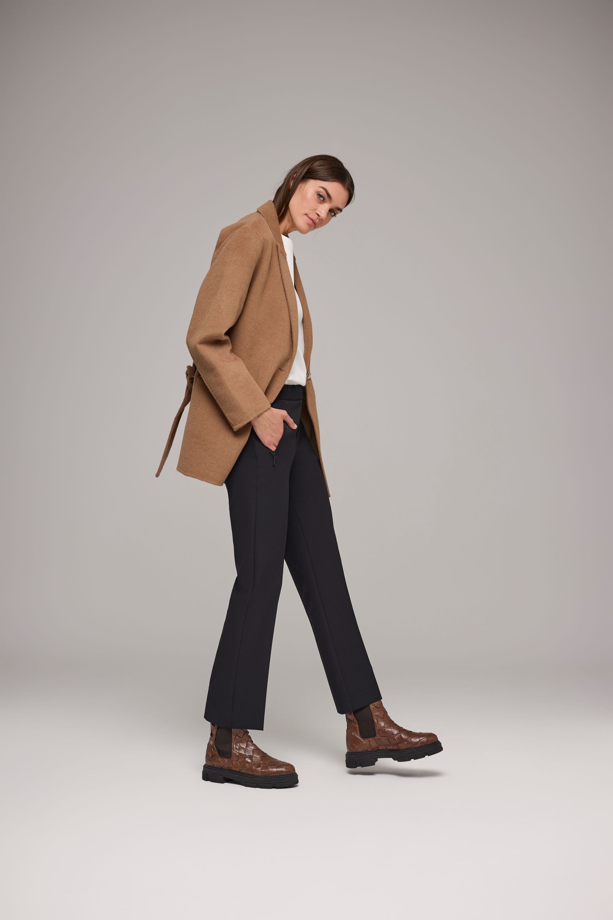 Frau trägt schwarze, leicht ausgestellte Hose mit Gummibund, kombiniert mit weißem Oberteil, beigem Mantel und braunen Chelsea-Boots.