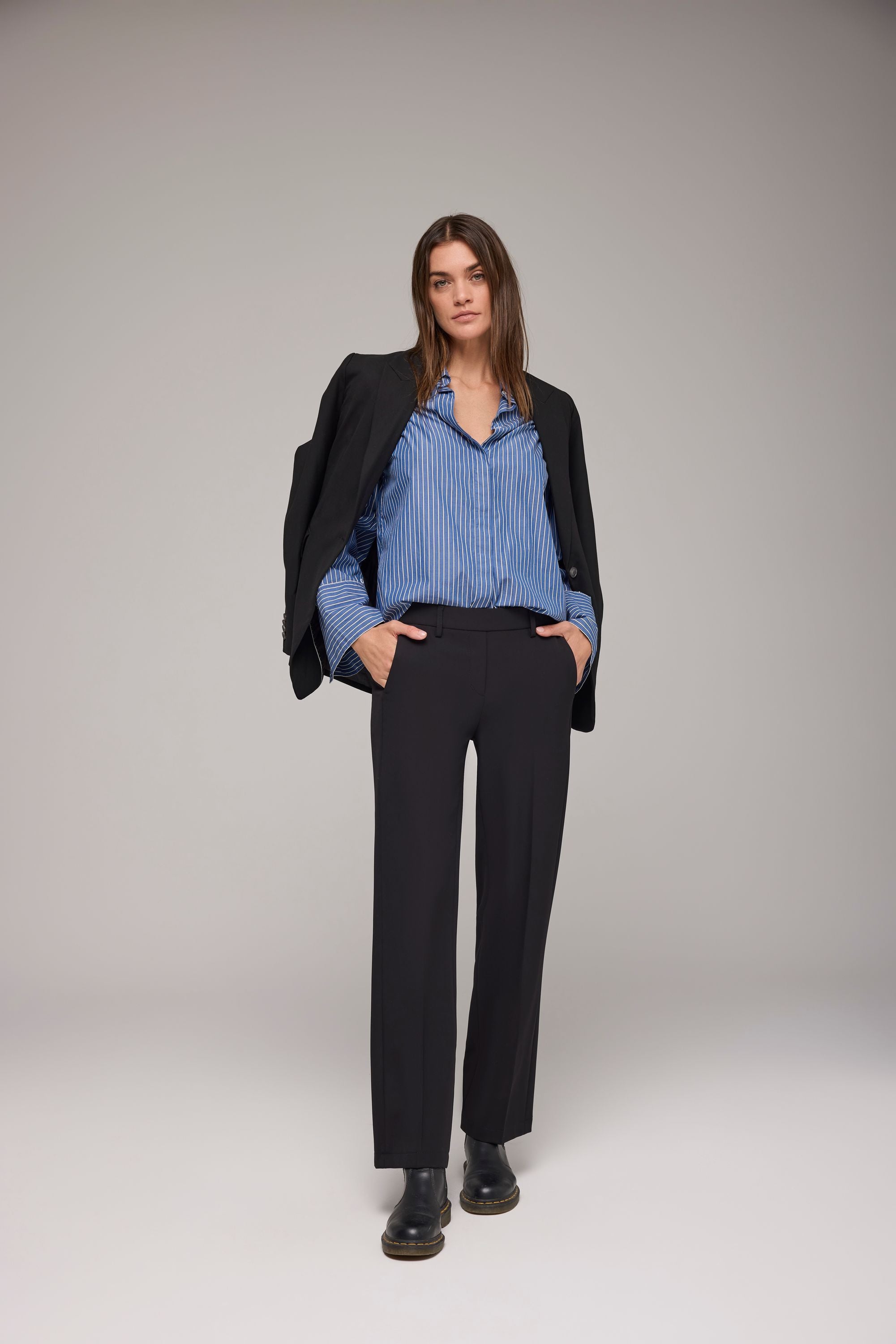 Frau trägt schwarze Stoffhose, graues Stricktop über gestreifter Bluse und schwarze Loafer – stehend auf Betonboden vor weißer Wand.