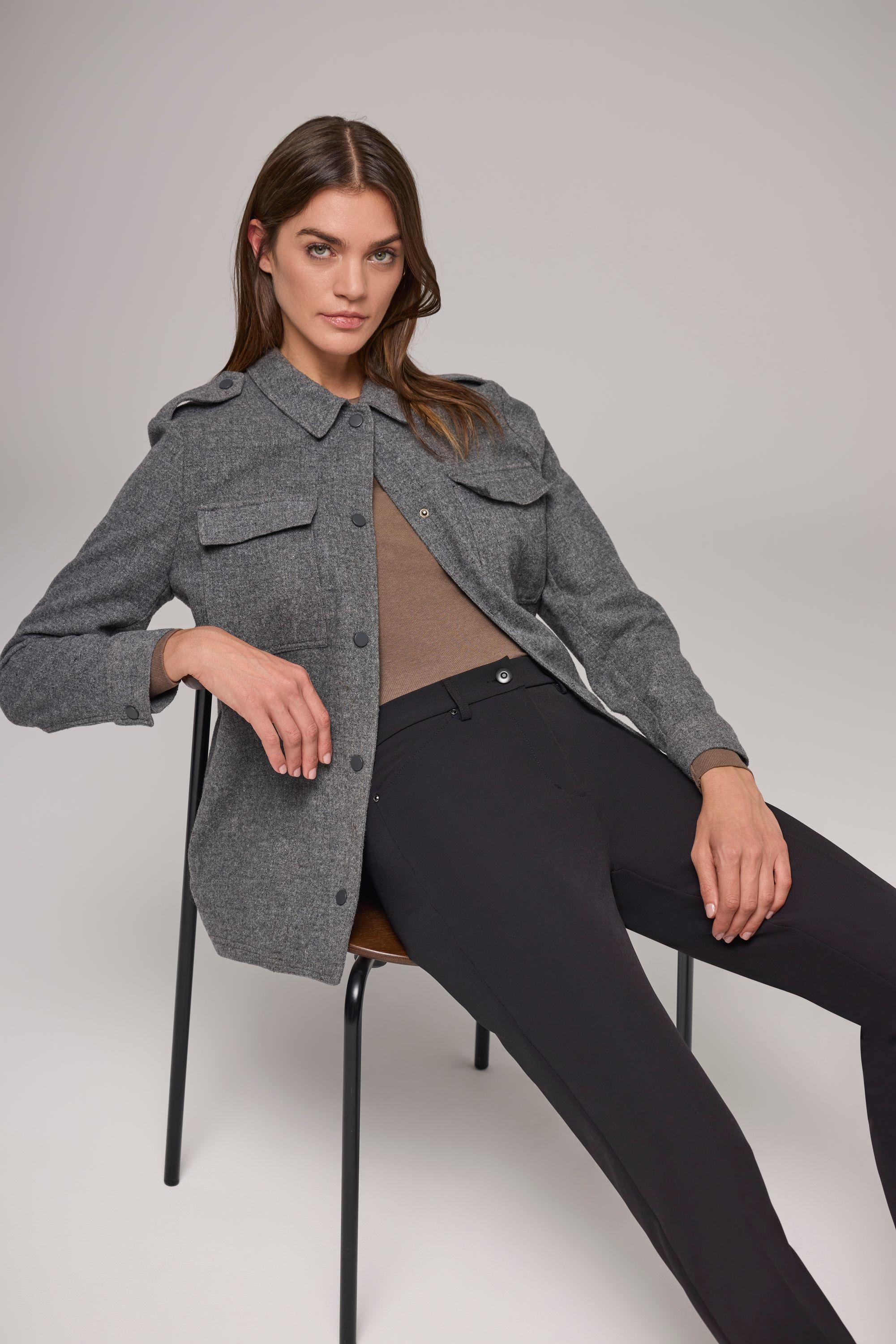 Frau sitzend auf Stuhl mit schwarzer Hose, braunem Top und grauer Jacke.